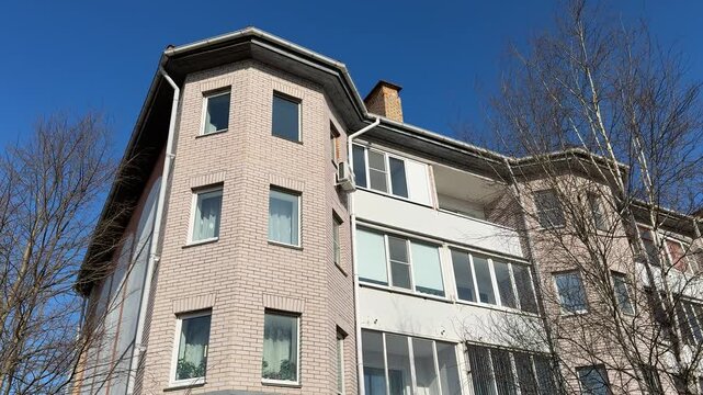 White brick apartment against blue sky. Sunlit multistorey facade with rows of windows, balconies and chimney framed by bare