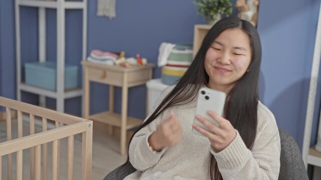 Young chinese woman smiling and holding a smartphone beside a wooden crib in a building nursery; motherhood calm bonding.
