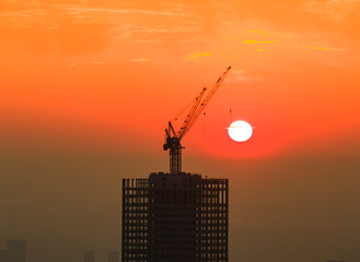 Silhouette of a skyscraper under construction in Wuhan, China, during a bright orange sunrise. A towering crane perfectly frames the glowing sun, symbolizing urban development and real estate. © 晨啸 周
