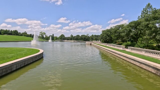 Water fountains spray and blow in the wind inside a large public park basin, bordered by an ornate stone banister and grassy hillside at Forest Park in St. Louis, Missouri, USA.