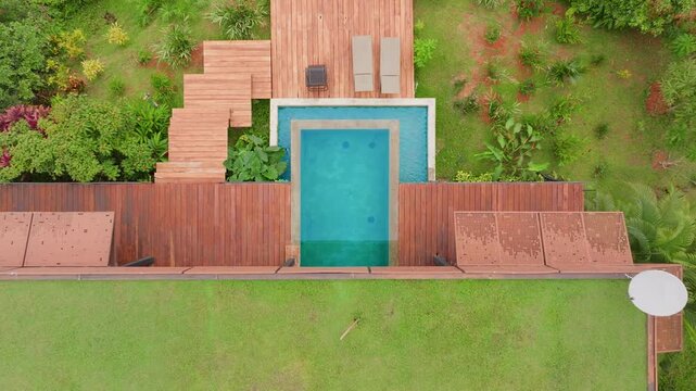 Aerial drone footage reveals stunning luxury villa in Costa Rica featuring turquoise infinity pool with wooden deck terraces surrounded by lush tropical gardens and manicured lawn.