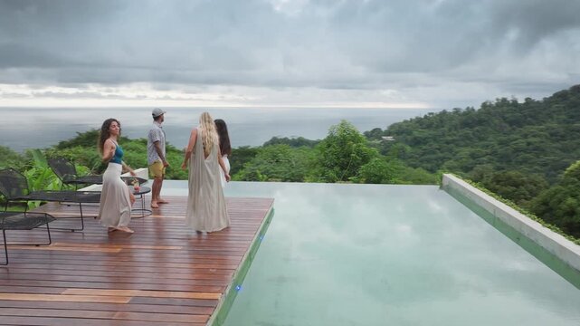 Group of young people dancing by pool at a small party on a wooden deck overlooking lush tropical jungle and ocean in slow motion. Friends celebrate together at luxury villa with dramatic cloudy sky.