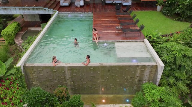 Group of young people dancing by pool at a small party. Slow motion aerial view captures friends enjoying luxurious infinity pool surrounded by lush tropical garden with wooden deck and modern villa.