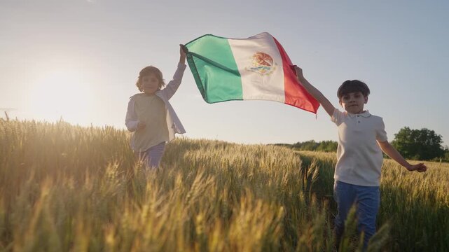 Little children joyfully run through country road waving the Mexican flag, flutters in the warm breeze above yellow wheat fields. Latin American pride and freedom.