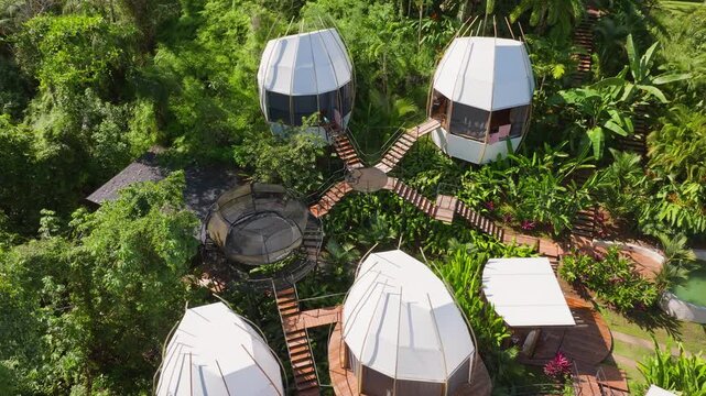Aerial drone footage reveals futuristic pod-shaped glamping accommodations suspended above lush tropical rainforest canopy, connected by wooden walkways and staircases amid vibrant green foliage.