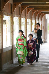Fototapeta premium Family walks under arched wooden corridor. Mother holds basket, father carries child. Daughters in colorful kimonos smile gently. Soft light filters through pillars behind them