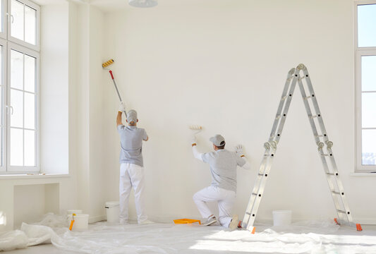 Team of professional painters in uniform painting white wall in empty room with paint rollers. Building contractors doing renovation in client house. Repair service and renovating concept.