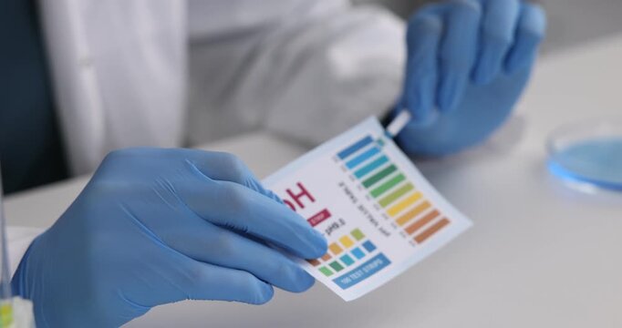 Scientist holding PH test strip with color table at white table in laboratory, closeup