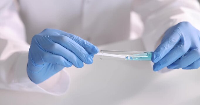 Scientist holding PH test strip and glass tube with liquid at white table in laboratory, closeup