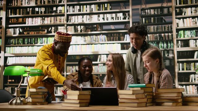 Diverse students working on laptop together at group study session, peers and adult learner gathering at library table with books and study materials.