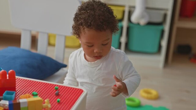 Toddler girl in white shirt holding a blue ball and reaching with hand while playing by a toy table and colorful play mat inside building; curiosity.