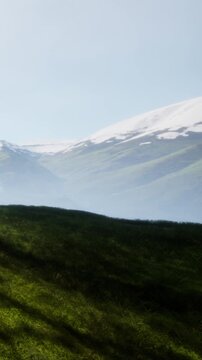 Mountains stand tall in the distance with snow on the peaks. Green fields stretch in the foreground. The sky is clear and bright.