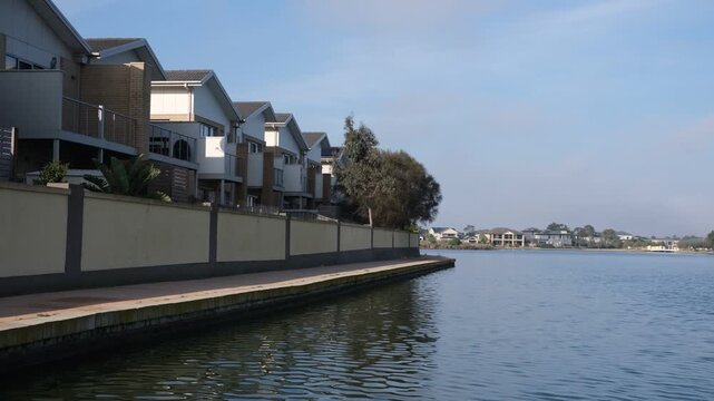 Modern waterfront townhouses line a boardwalk beside a calm suburban lake in Sanctuary Lakes, Point Cook, Melbourne Australia.&nbsp;Contemporary Australian waterside living.