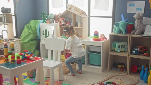 Girl carrying wicker basket of dolls and toys across playroom in building with wooden train track and storage shelves; playful exploration.