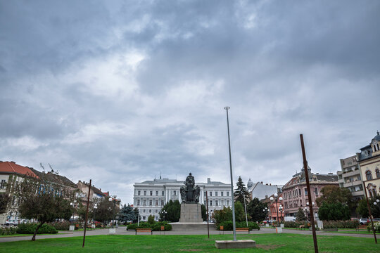 Avram Iancu Square in Arad, Romania, with the Ioan Slavici Theater behind a central monument, lawns, benches and historic buildings under a cloudy sky in the city center of Northern Romania.