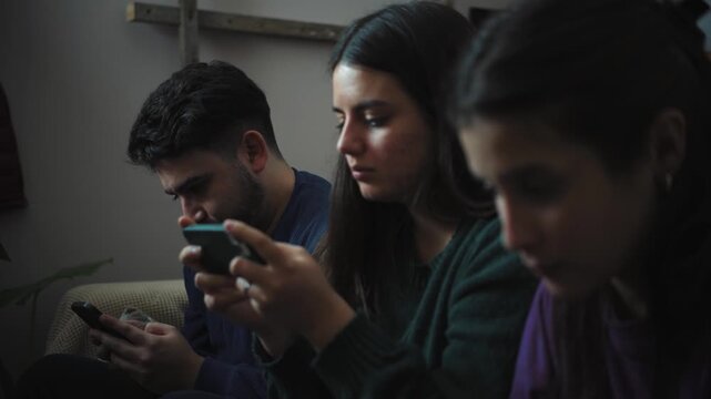 Group of young Hispanic friends sitting on a sofa, each absorbed in their own smartphone instead of socializing, highlighting digital distraction, technology dependence, and the impact of mobile devic