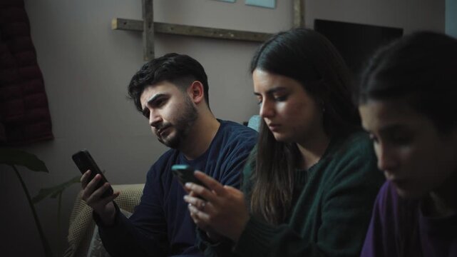 Three young Hispanic friends sitting on a sofa looking bored while using their smartphones, occasionally glancing at the TV, illustrating digital distraction, multitasking, and a casual evening at hom
