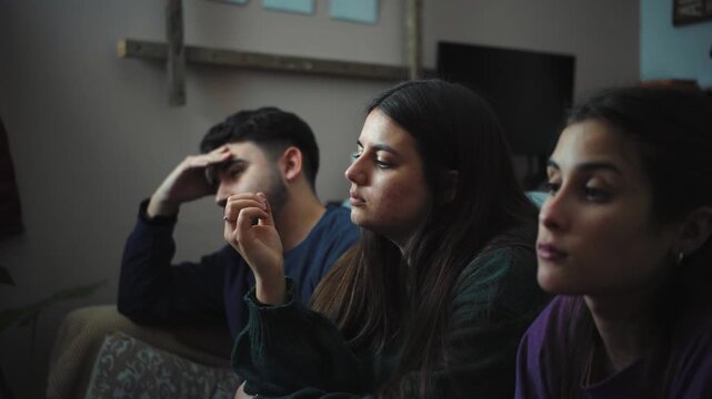 Three young Hispanic friends sitting on a sofa sharing an awkward moment while watching a movie on TV, showing boredom, silence, and a lack of interest in the film during a quiet evening at home.

