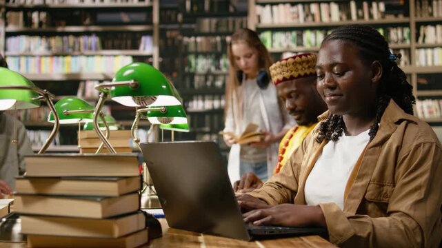 Black woman student uses laptop at shared library table with books and green lamps, surrounded by many study materials while peers read and work nearby, for online learning, research and coursework.