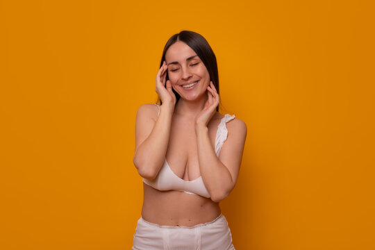 Smiling young woman touching healthy facial skin with closed eyes on orange background