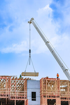 Low angle view of a construction crane telescoping boom lifting a large bundle of lumber framing materials to workers on top of a residential building site