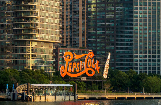 New York, NY - US - Jul 1, 2026 Iconic Pepsi-Cola sign glowing in warm sunset light along Long Island City waterfront, framed by modern glass towers and calm East River reflections.