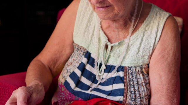 Elderly woman with a santa hat carefully sewing a handmade christmas decoration, using a needle and thread to create a unique felt ornament for the holiday season while sitting on a red armchair
