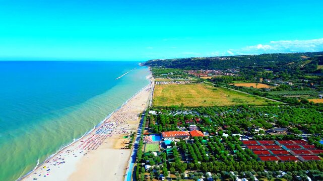 Delicate aerial push-in shot from Marina Palmense with the vast rippling waters of the Adriatic Sea skimming the sandy beach and creating creamy foam, with parasols, roads, trees, buildings, hillscape