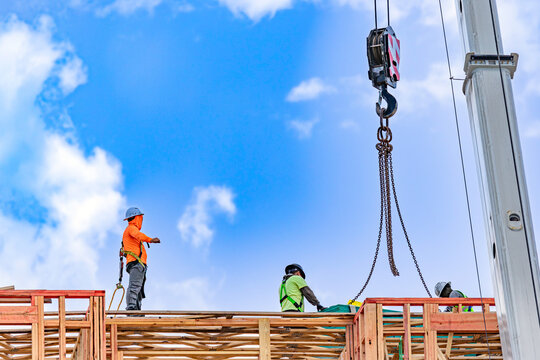 Low angle view of construction workers in safety harnesses and hard hats guiding a crane lifting hook with chains on top of a residential building wood framing structure