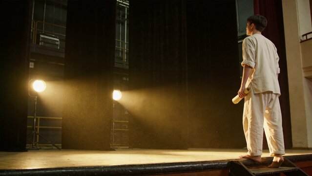 Actor on Stage: A lone actor stands on the edge of a dimly lit stage, illuminated by two bright spotlights, holding a scroll, ready to perform.