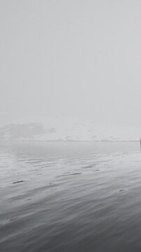 A person in a small boat navigates through cold waters with fog covering the landscape. Snow can be seen in the distance. The scene shows winter life on the water.