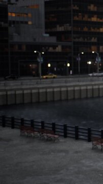 People are walking near the river as city lights reflect on the water. Benches line the riverbank, and buildings stand tall in the background during the evening.