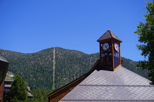 A scenic view of a rustic wooden building with a prominent four-sided clock tower and a grey shingled roof. In the background, a massive mountain covered in dense green pine trees rises up under a bri