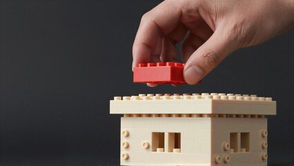 Fototapeta premium Hand places red LEGO brick atop beige structure, symbolizing growth or construction.
