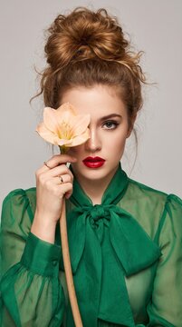 Elegant woman with messy bun hairstyle holding a soft peach amaryllis flower over her eye while wearing a vibrant green sheer blouse in studio light