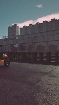 A tractor is parked on a wet surface next to a tall building. The large wall has figures painted on it. The scene takes place in an industrial area during daytime.