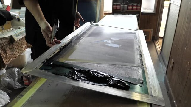 Standing artisan drags a large silkscreen squeegee across a long fabric panel spreading black dye