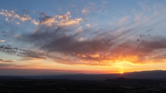 4K UHD drone timelapse capturing a vibrant sunrise over a dark Southwestern canyon landscape. Wispy clouds dramatically change colors, radiating bright golden and orange hues as the sun ascends.