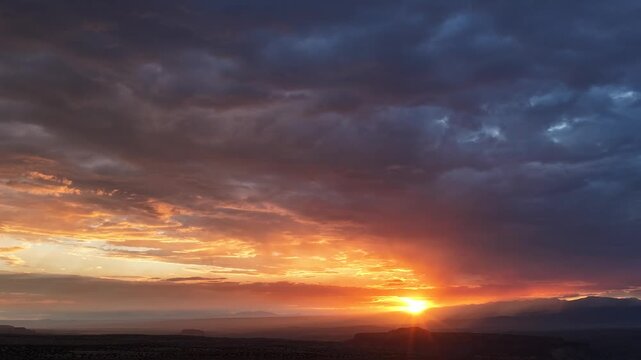 4K UHD drone timelapse of a stunning sunrise cresting a dark mountain silhouette. Intense golden light ripples across heavy clouds, radiating vibrant colors over the vast desert landscape below.