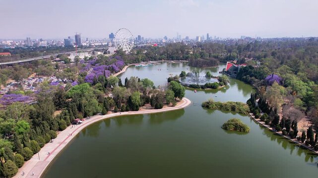 Drone flight over the lake in Chapultepec Second Section featuring the Aztl&aacute;n Ferris wheel, fountains, jacaranda trees and the Mexico City skyline on a clear day.