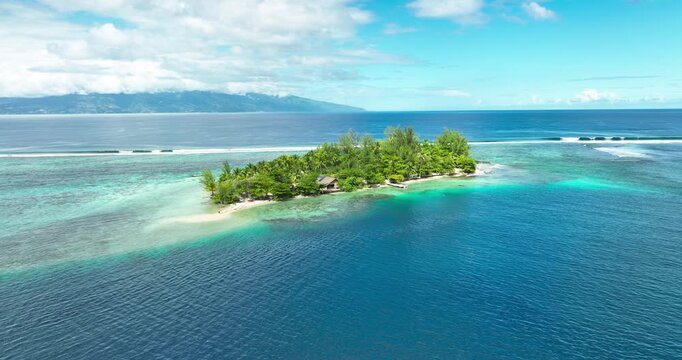Tiny palm-covered motu sits isolated amid vast turquoise lagoon, surrounding coral reef fading into deep blue Pacific waters near distant French Polynesian mountains. Aerial drone wide shot