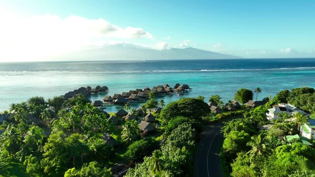 Overwater bungalows line turquoise lagoon beside lush Moorea coastline, curving road winding through palm trees toward misty island mountains under bright Pacific sky. Aerial drone wide shot