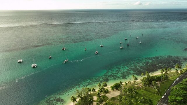 Dramatic jagged peaks rise above Ta&rsquo;ahiamanu Beach coastline in Moorea, coastal road winding past village houses toward calm turquoise bay dotted with anchored boats. Aerial drone wide shot
