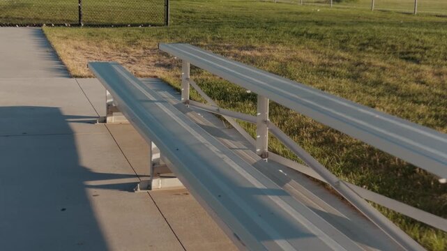Camera glides over and past an empty bleacher bench beside a sports field
