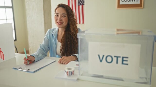 Woman writing on clipboard with pen at polling station beside a clear ballot box and american flag, smiling at camera; civic duty pride.