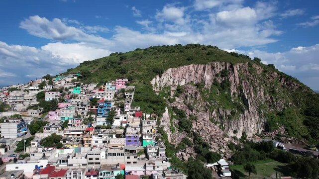 Drone flies toward Cerro Gordo in Ecatepec, revealing the rocky cliff, colorful hillside homes and lush vegetation beneath a bright blue sky with dramatic clouds.