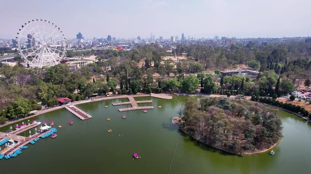 Drone flies toward Chapultepec Second Section lake, showcasing the Aztl&aacute;n Ferris wheel, island, paddle boats and surrounding park in Mexico City on a sunny day.
