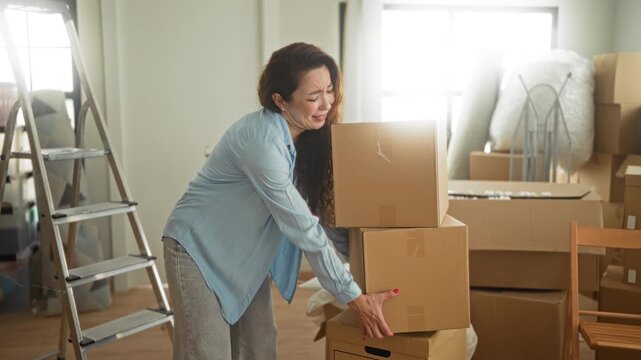 Woman clutching lower back with backache while lifting cardboard boxes in a building; moving day stress pain.