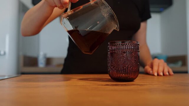 A woman pours hot filtered coffee from a glass V60 server into a textured drinking glass on a wooden kitchen counter.