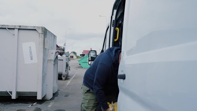 Man shuts the side door of a white van, then picks up a heavy rubbish bag at an outdoor waste management center.